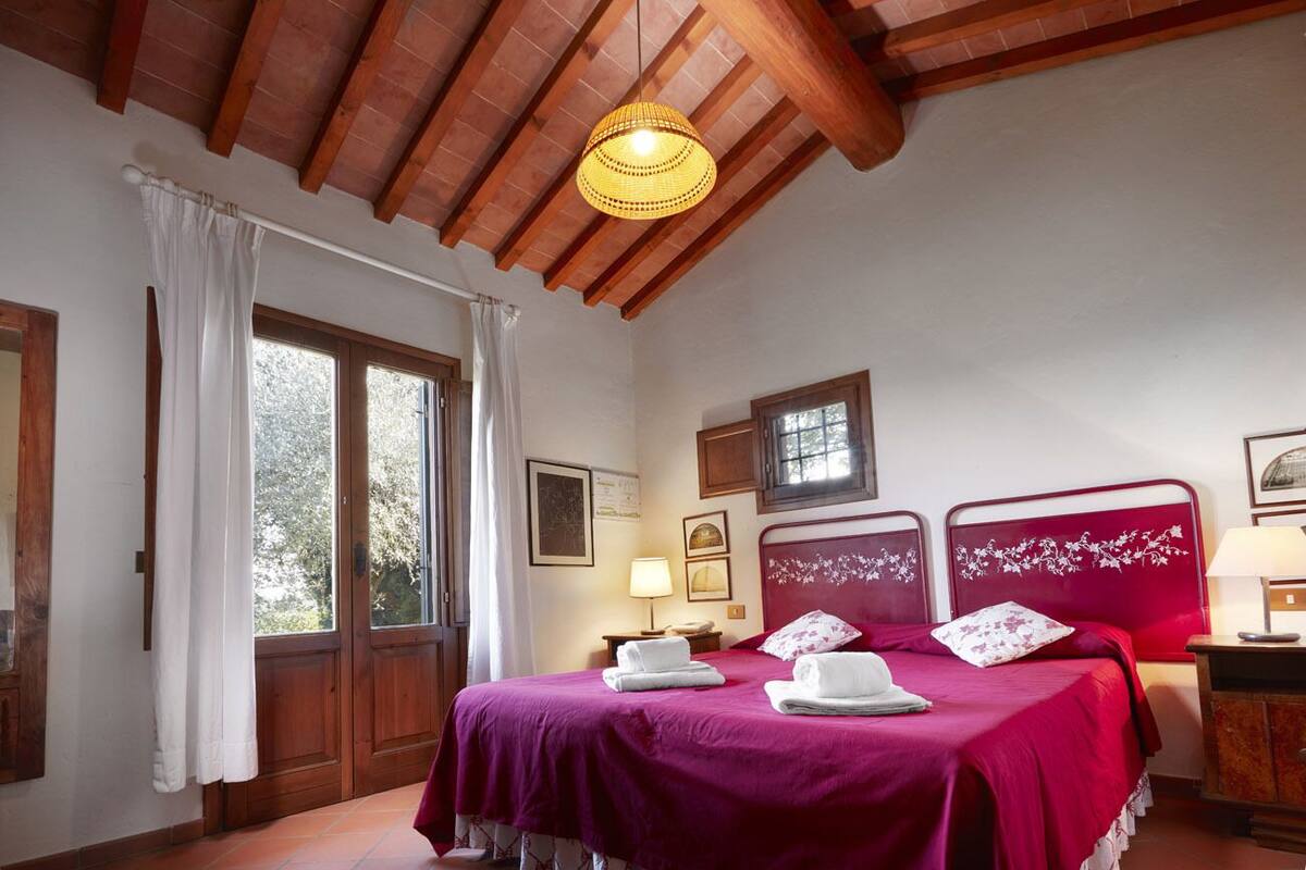 Une chambre avec deux lits, des poutres en bois au plafond, une fenêtre avec des rideaux et une table de chevet avec une lampe.