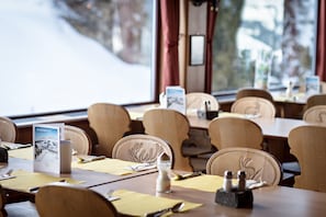 A dining area with wooden tables and chairs, a window with a snowy view, and a menu stand with a book titled "TRÜBSEEDPOST".