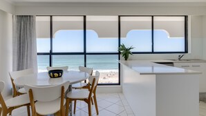Una cocina moderna con un ventanal grande con vistas a la playa, un comedor con sillas blancas y una mesa, y una pequeña isla con un lavamanos.