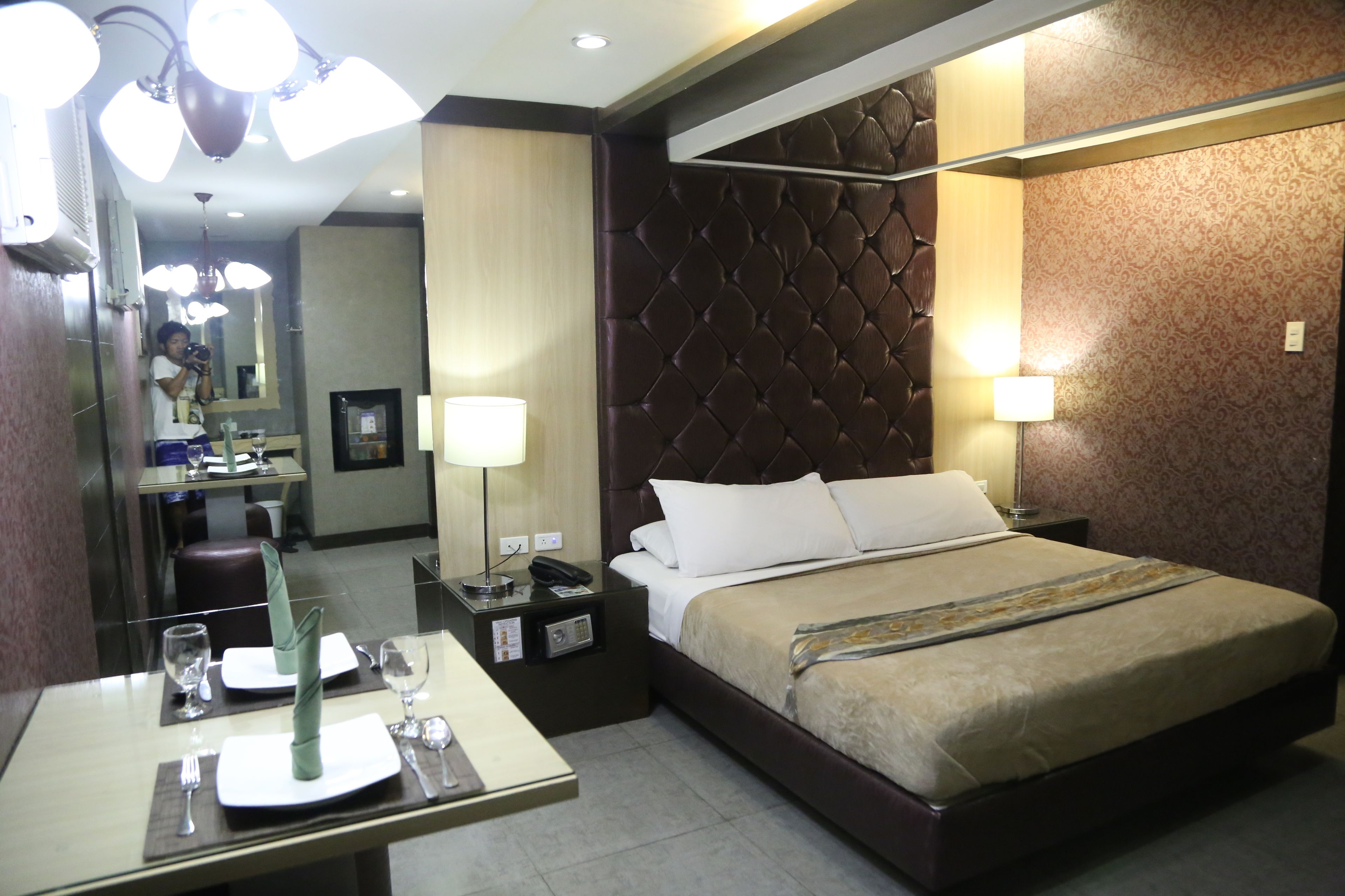 Une chambre d’hôtel avec un lit soigneusement fait, une table de chevet avec une lampe, une petite table de nuit avec un téléphone, et un coin repas avec une table mise pour deux personnes.