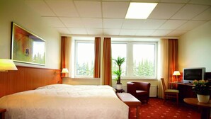 Una camera d'albergo con un letto grande, una scrivania con televisione, una sedia, un tavolino con una pianta e una finestra con tende.