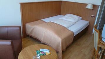 Habitación de hotel con cabecera de madera, una cama individual con colchón blanco y manta marrón, una mesita redonda pequeña con un vaso y un control remoto, y una silla de cuero marrón.