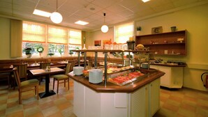 Un'area buffet con diversi tavoli e sedie da pranzo, un banco centrale per il servizio con varie pietanze, e una cucina sullo sfondo.