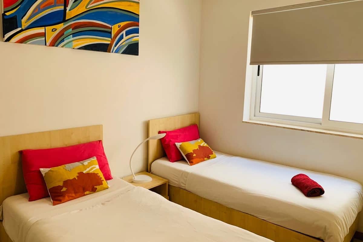 A room with two single beds, each with a wooden headboard and white bedding. There is a colorful abstract painting on the wall above the beds, and a window with a white blind to the right.