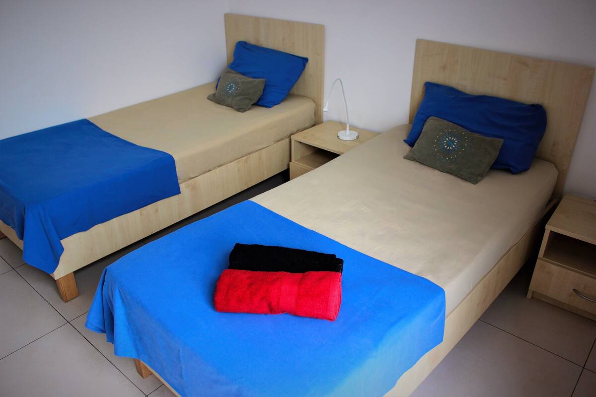 Two single beds with wooden headboards, a nightstand with a lamp, and a red and black object on the bed.