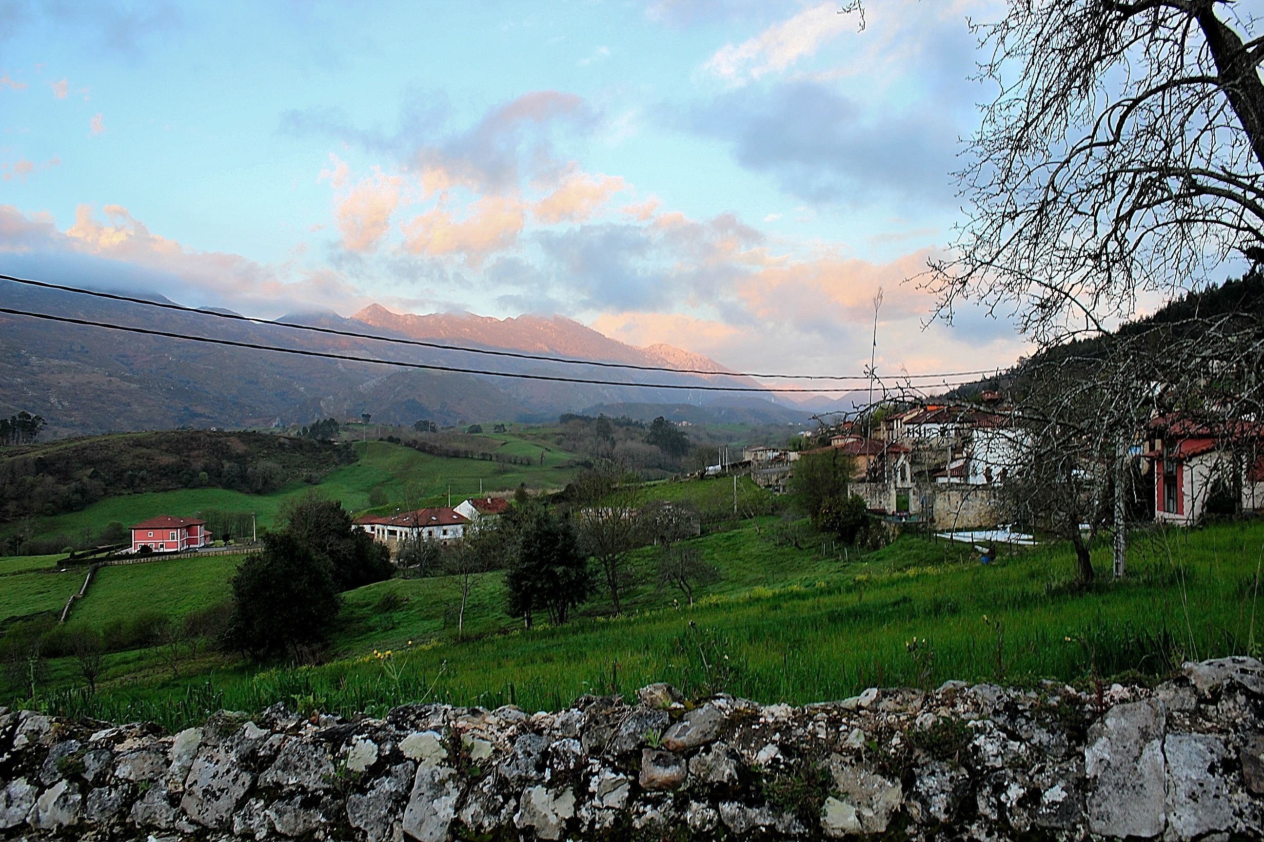Un paysage rural avec des maisons, un mur de pierre et une chaîne de montagnes en arrière-plan.