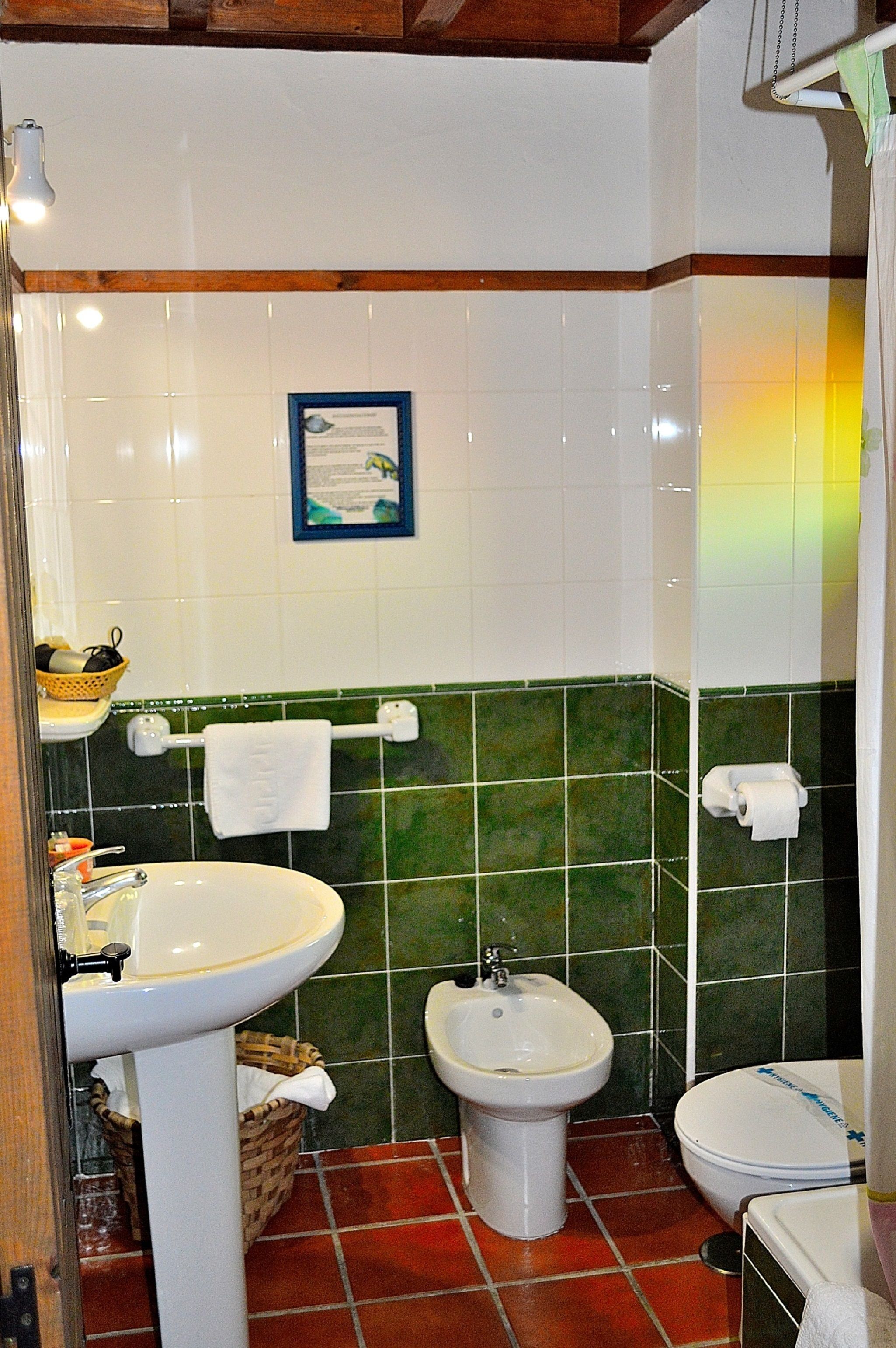 A bathroom with a sink, bidet, and toilet. There is a towel rack with a towel, a basket, and a framed certificate on the wall.