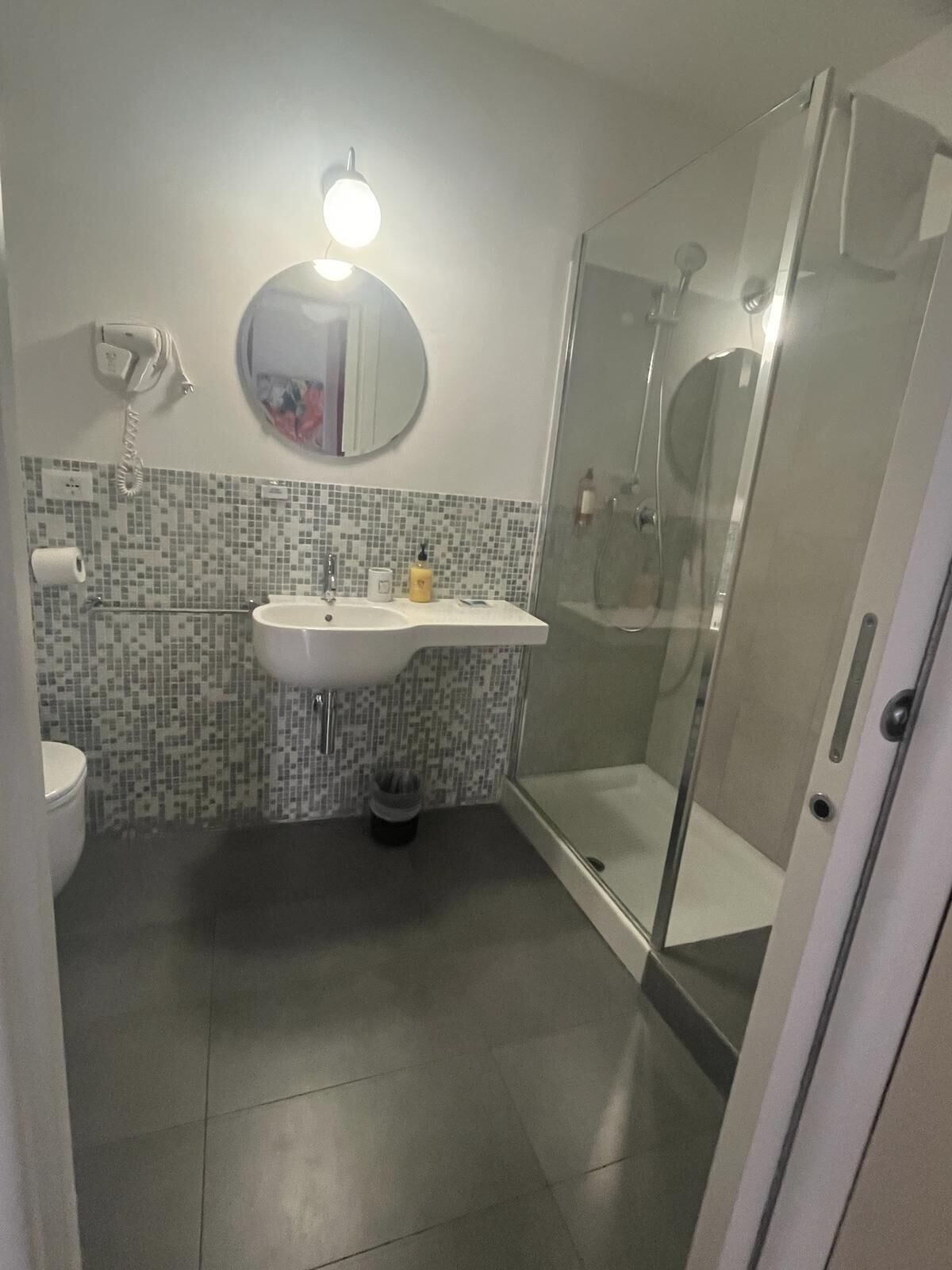 Un baño con piso de baldosas, un lavamanos con espejo, un inodoro y una ducha con mampara de vidrio.