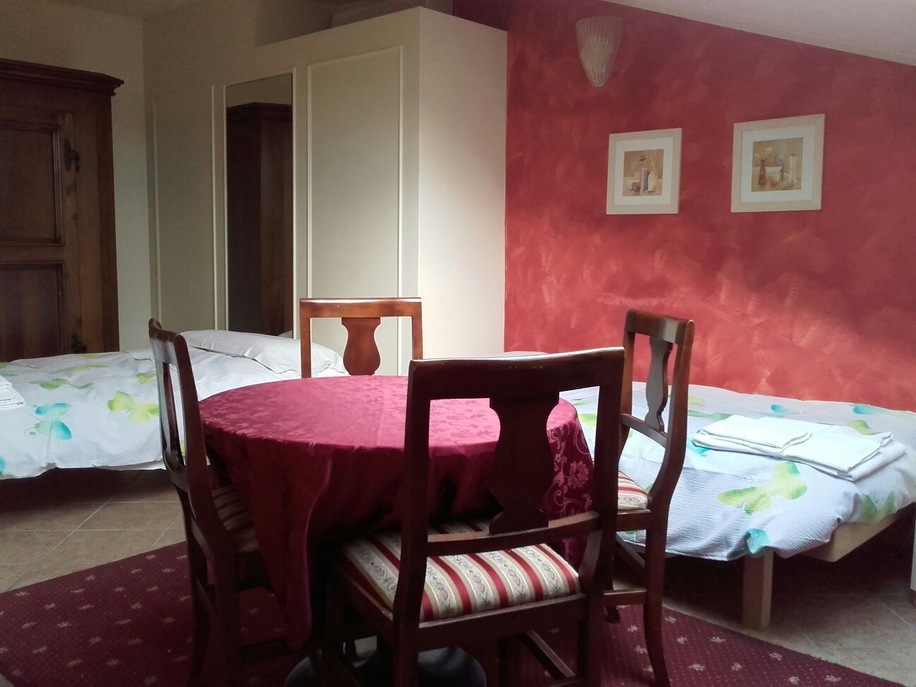 A room with a round table and four chairs, two beds with floral bedding, and a red accent wall with framed pictures.