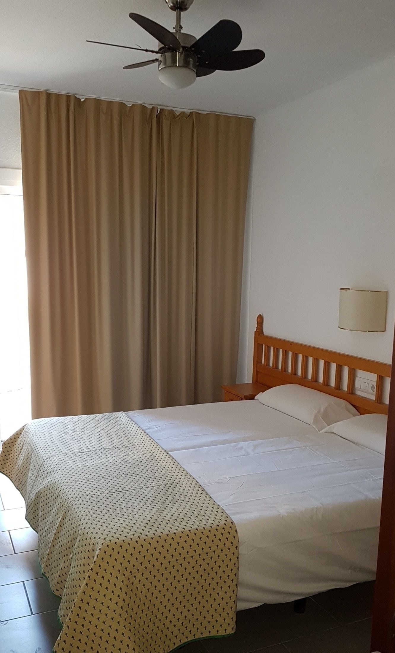 A single bed with a wooden headboard, a ceiling fan, and a window with curtains.