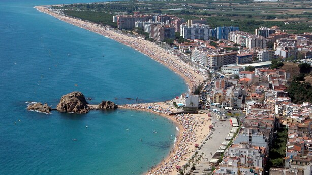 Un paisaje urbano costero con una playa extensa, edificios urbanos densos y un promontorio rocoso cerca de la orilla.