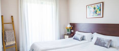 Habitación de hotel con dos camas, una escalera de madera, una mesita de noche con lámpara y flores, y un cuadro en la pared.