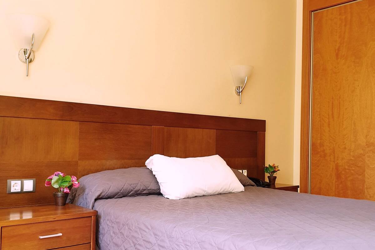 A neatly made bed with a quilted cover, a wooden nightstand with drawers, a wall-mounted lamp, and a wooden wardrobe.