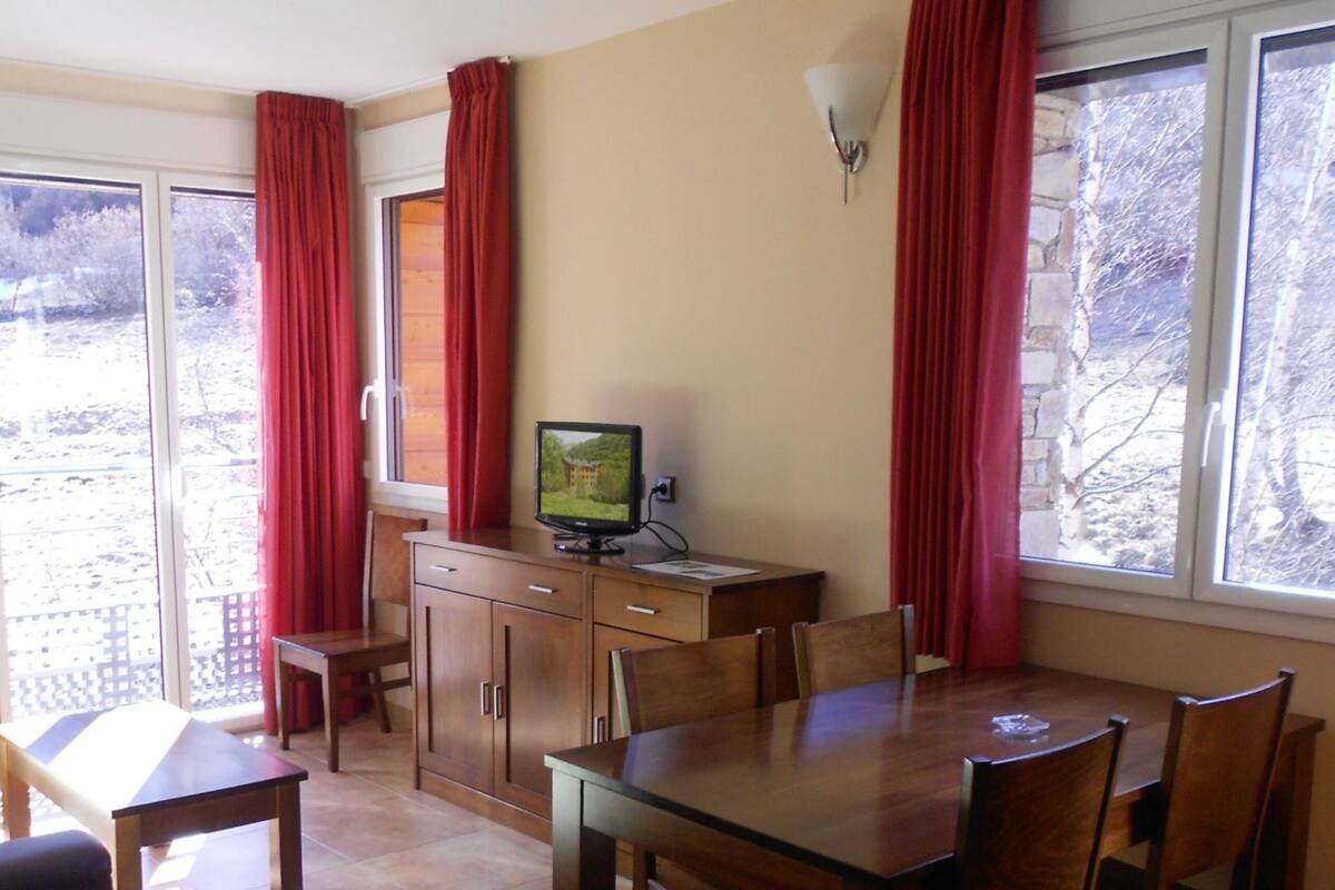 A dining area with a wooden table and chairs, a television on a cabinet, and large windows with red curtains.