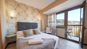 A hotel room with a large bed, a small table, a chair, and a balcony with a view of the hills. - Hotel Sánchez (Ainsa)