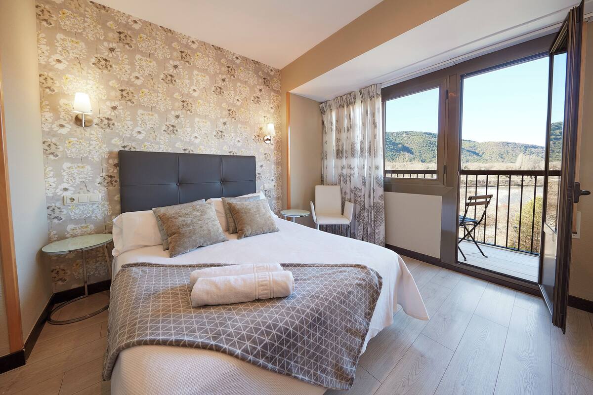 A hotel room with a large bed, a small table, a chair, and a balcony with a view of the hills.