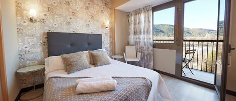 Habitación de hotel con una cama grande, una mesita, una silla y un balcón con vistas a las colinas.