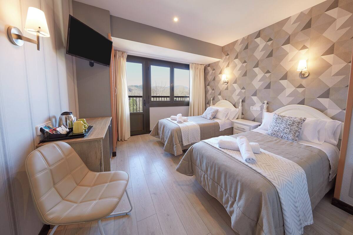 A hotel room with two beds, a TV, a desk with a chair, and a view of the outdoors.
