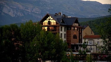 Ein mehrstöckiges Gebäude mit einem markanten Balkon, umgeben von Bäumen und Bergen im Hintergrund.