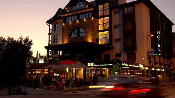 A hotel with a modern design, featuring a prominent 'HOTEL' sign, outdoor seating area, and multiple storefronts on the ground floor. - Hotel Sánchez (Ainsa)