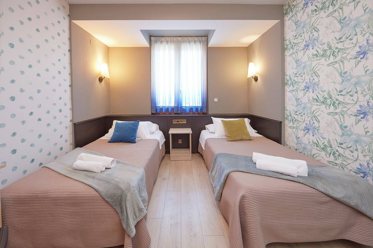 A hotel room with two beds, a small table, and a window with curtains.