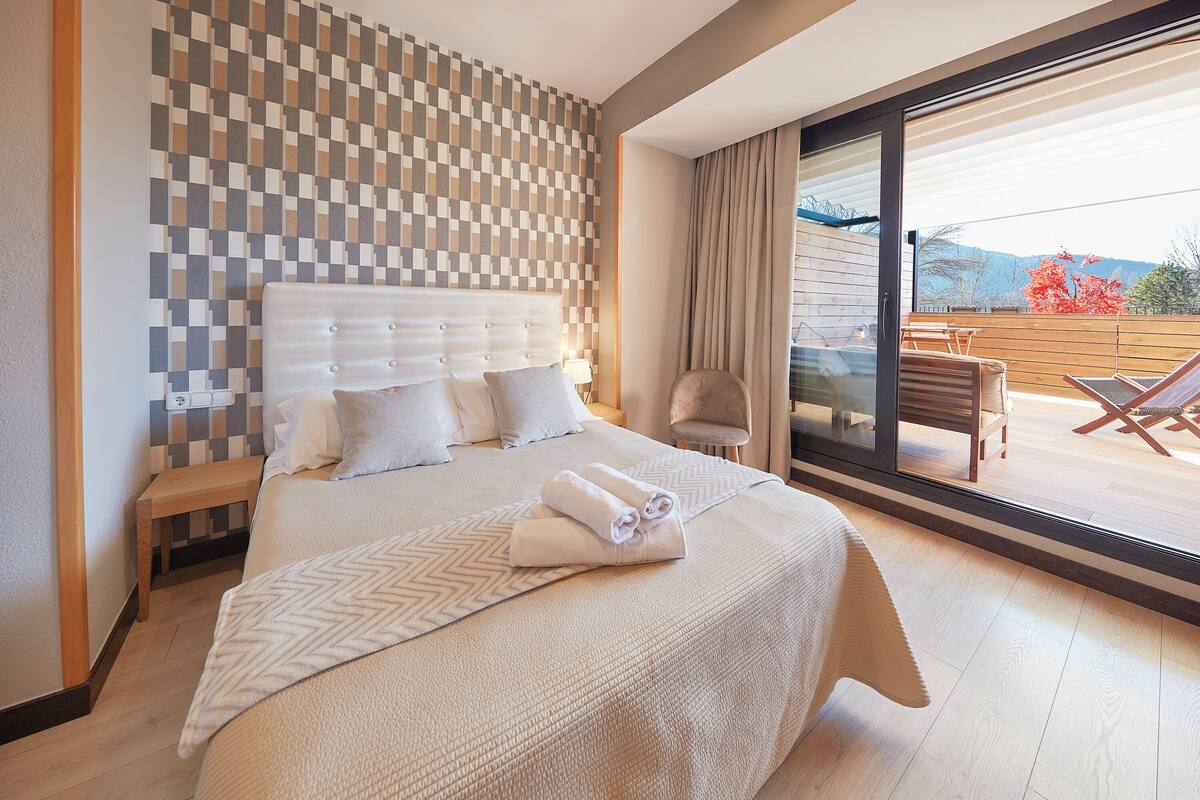 A modern hotel room with a large bed, a checkered wall, a balcony with a table and chairs, and a view of the outdoors.