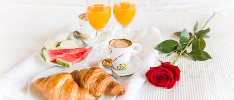 Desayuno con cruasanes, sandía, zumo de naranja, café y una rosa roja sobre un mantel blanco.