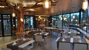 A restaurant with a modern interior, featuring a large mural, wooden tree-like structures, and hanging pendant lights. Tables are set with wine glasses and napkins. - Hotel Sánchez (Ainsa)