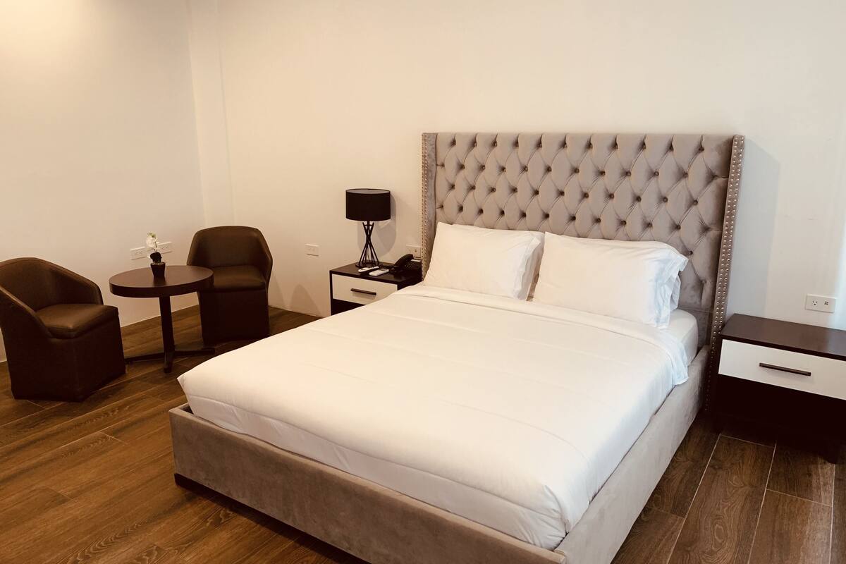 A hotel room with a large bed, a bedside table with a lamp, a chair, and a small table with a flower.