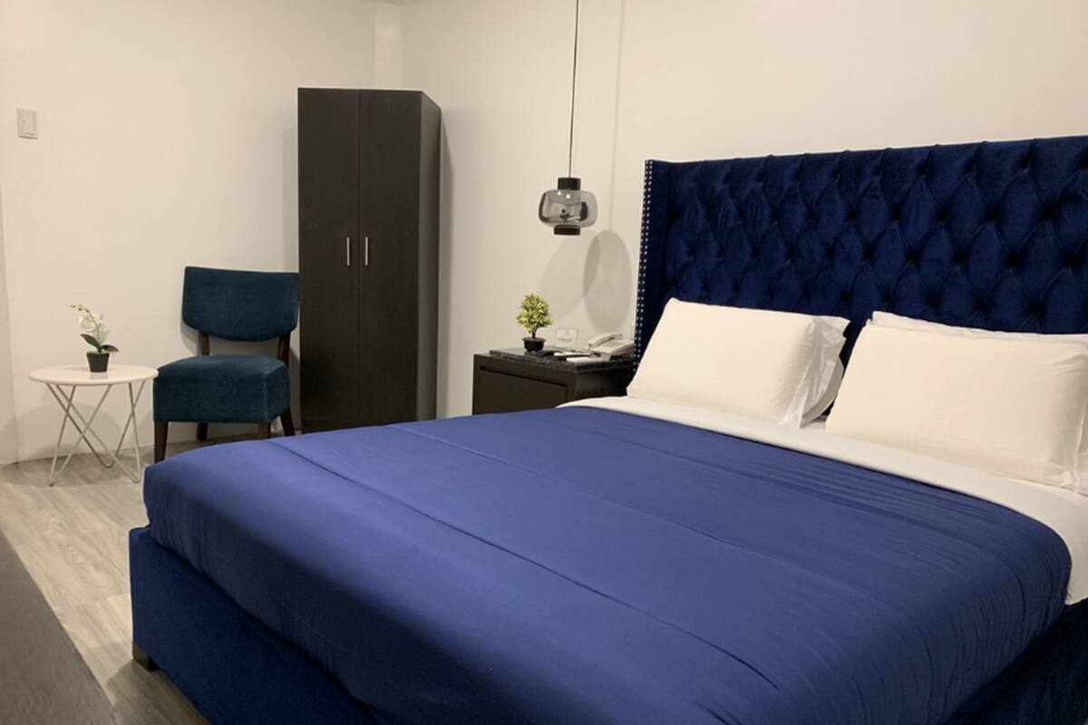 A bedroom with a large bed, a dark blue headboard, a wardrobe, a chair, a small table with a plant, and a nightstand with a lamp.