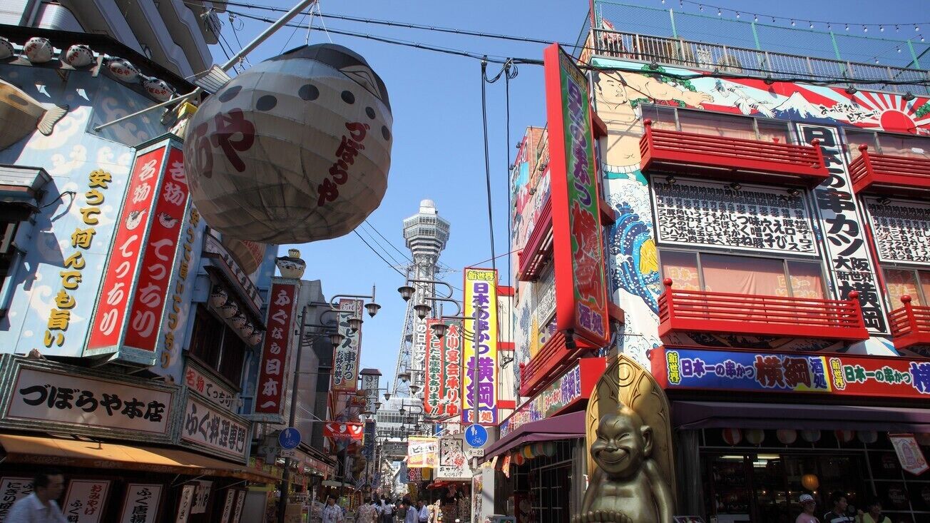 Une rue animée, bordée de divers commerces et de nombreuses enseignes, dont une grande enseigne rouge et blanche avec des caractères japonais.