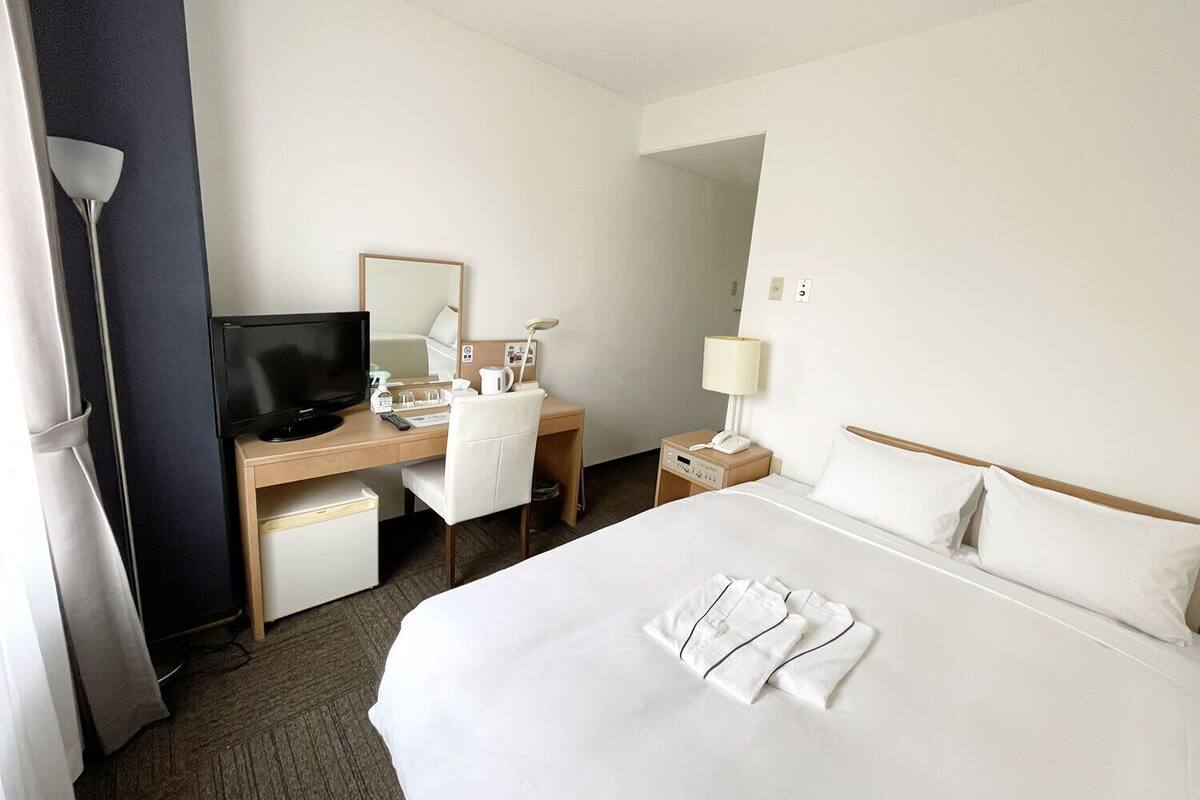 Una habitación de hotel con cama, escritorio con computadora, silla, lámpara de escritorio, teléfono y televisión.