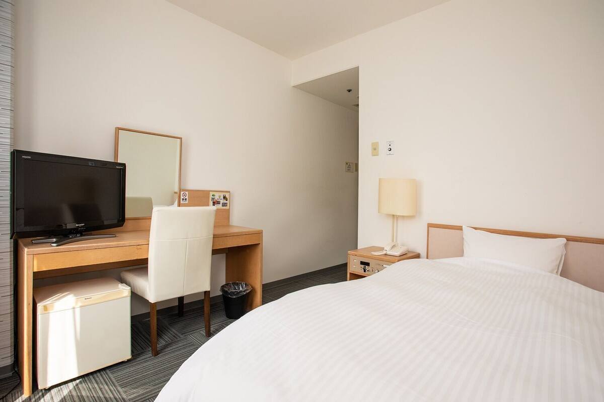 Habitación de hotel con cama, escritorio con televisor, silla, mesita de noche con lámpara y cubo de basura.
