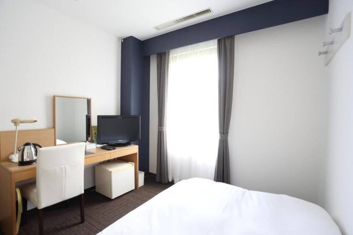Una habitación de hotel con cama, escritorio con computadora, silla, televisor y ventana con cortinas.