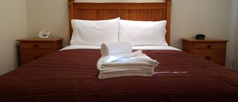 Habitación de hotel con cabecera de madera, dos almohadas blancas, una colcha roja y una pila de toallas blancas sobre la cama.