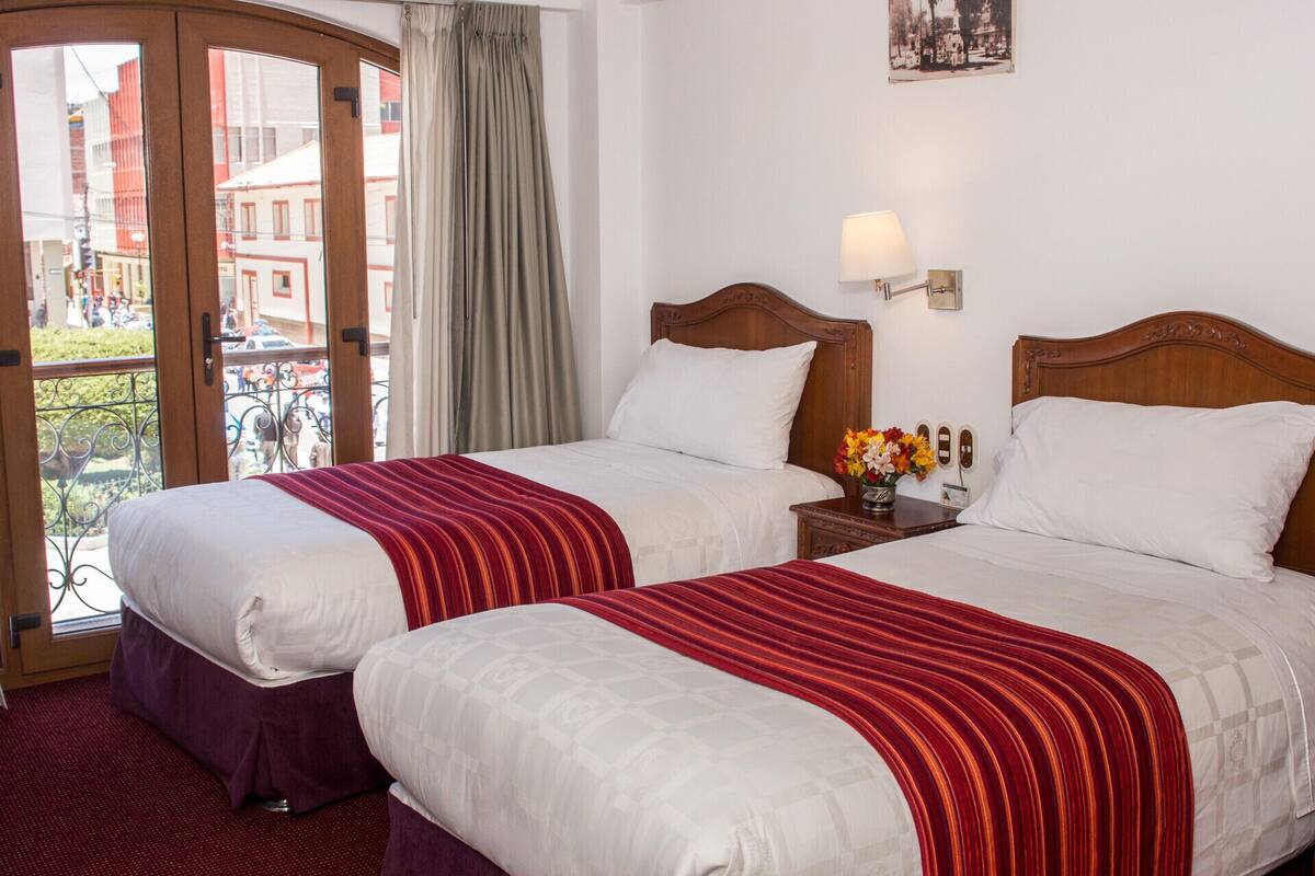 A hotel room with two beds, a balcony, a view of buildings, and a lamp on the nightstand.