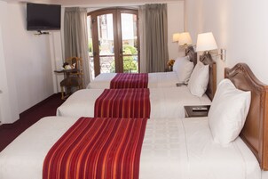 A hotel room with two beds, a TV mounted on the wall, a small table with a chair, and a view of the outside through a glass door.