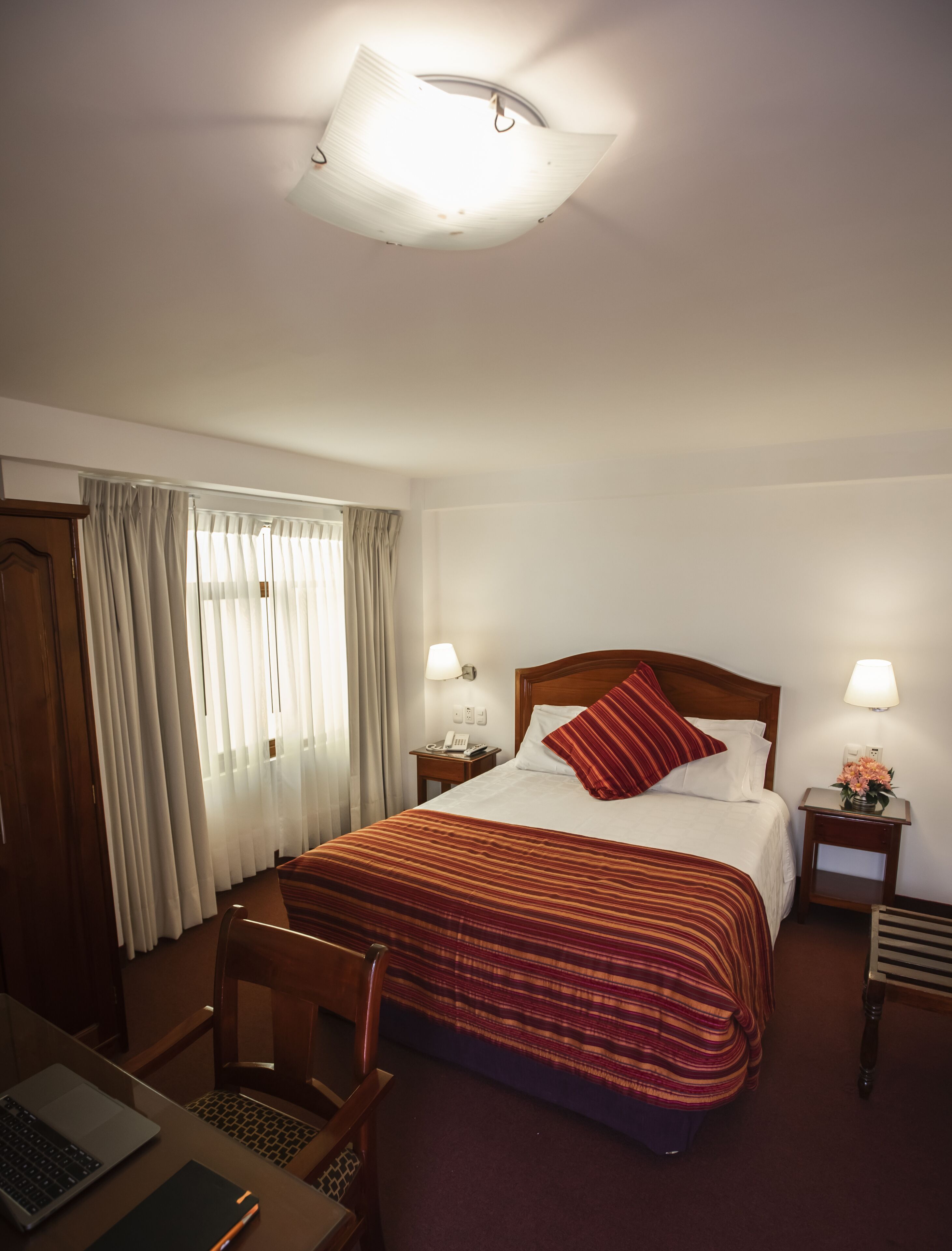 Une chambre d’hôtel comprenant un lit, un bureau avec un ordinateur portable, une chaise, une table de chevet avec une lampe et une fenêtre avec des rideaux.