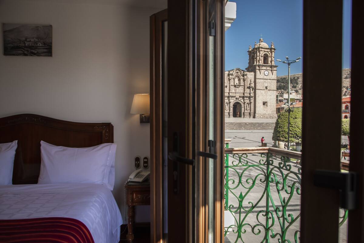 A hotel room with a bed, a bedside table with a lamp, and a view of a historic church through an open door.