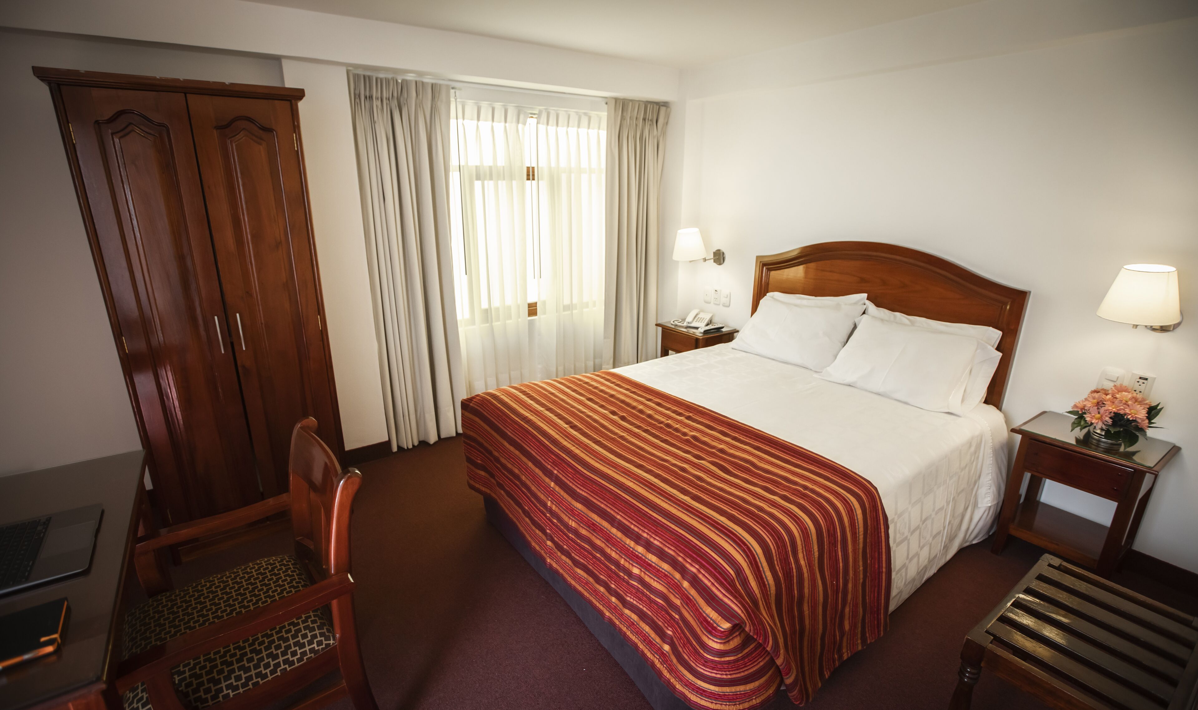A hotel room with a large bed, a desk with a chair, a wardrobe, and a bedside table with a lamp and flowers.