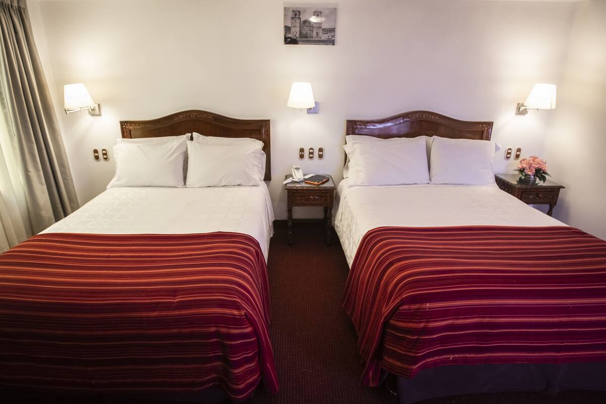 A hotel room with two beds, each with a wooden headboard, white bedding, and red striped bedspreads. There are bedside tables with lamps, a small wall-mounted picture, and a nightstand with a phone and a book.
