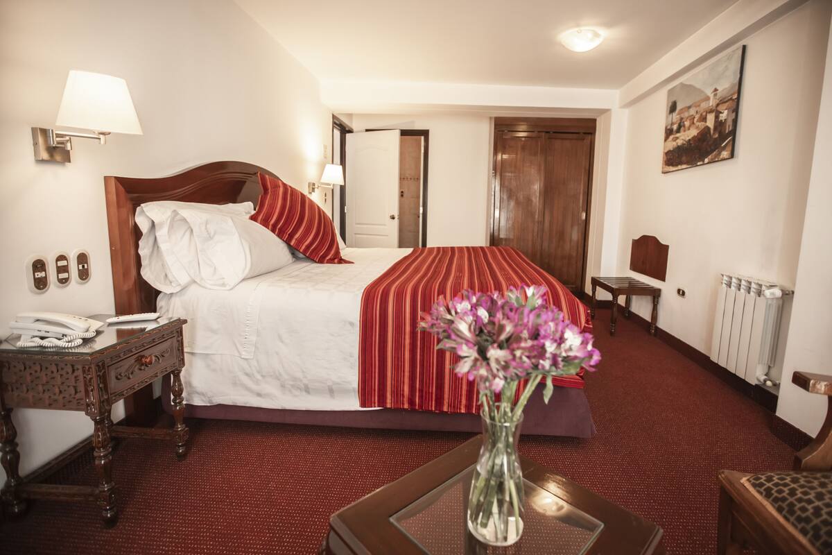 A hotel room with a bed, a bedside table with a lamp, a telephone, and a vase with flowers.