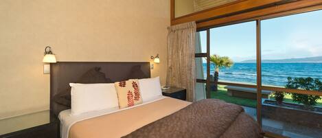 A bedroom with a large bed, a nightstand, a lamp, and a view of the sea through a large window.