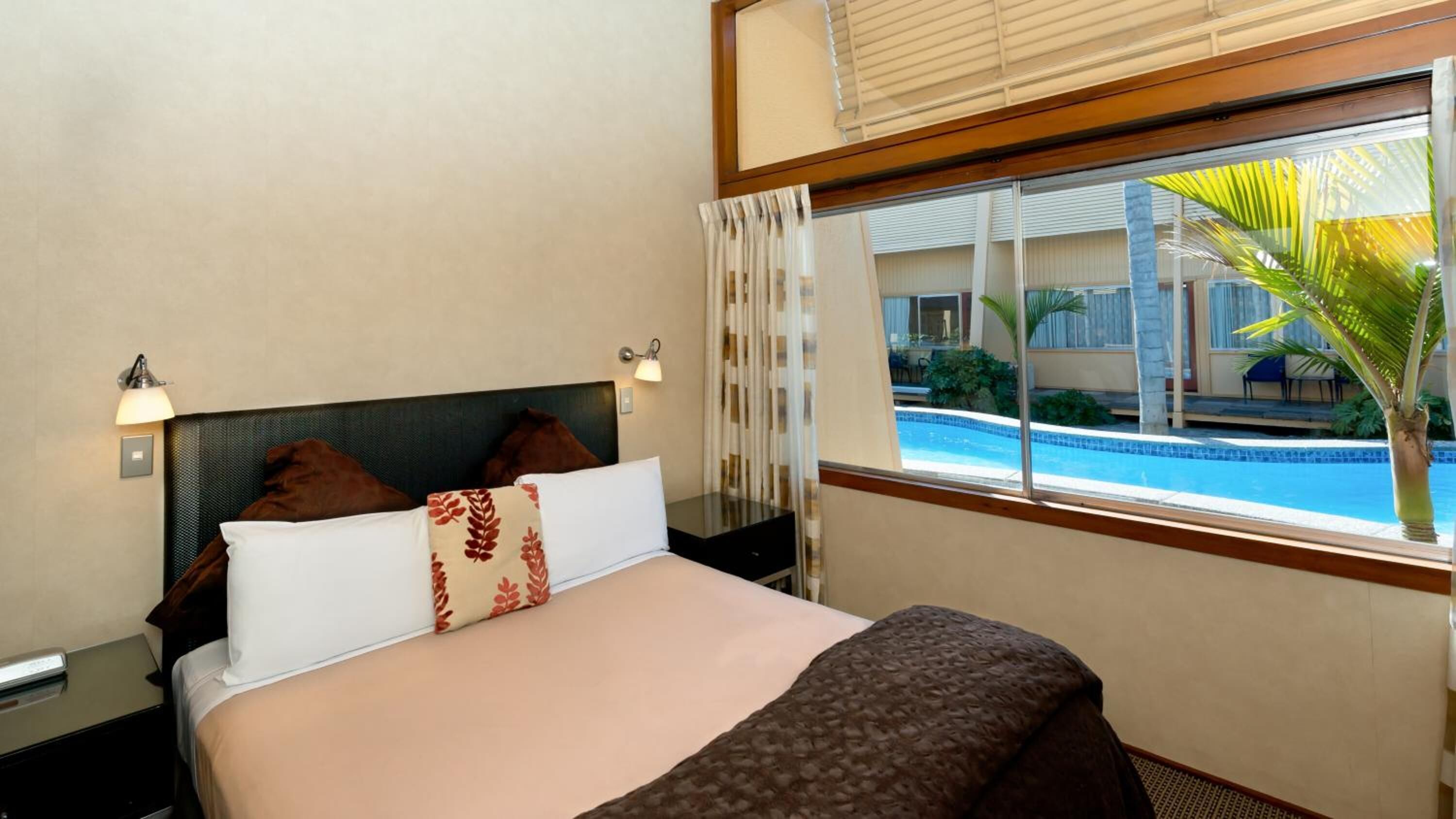 Une chambre d’hôtel avec un lit, une table de chevet, une lampe, une fenêtre donnant sur une piscine et des palmiers.