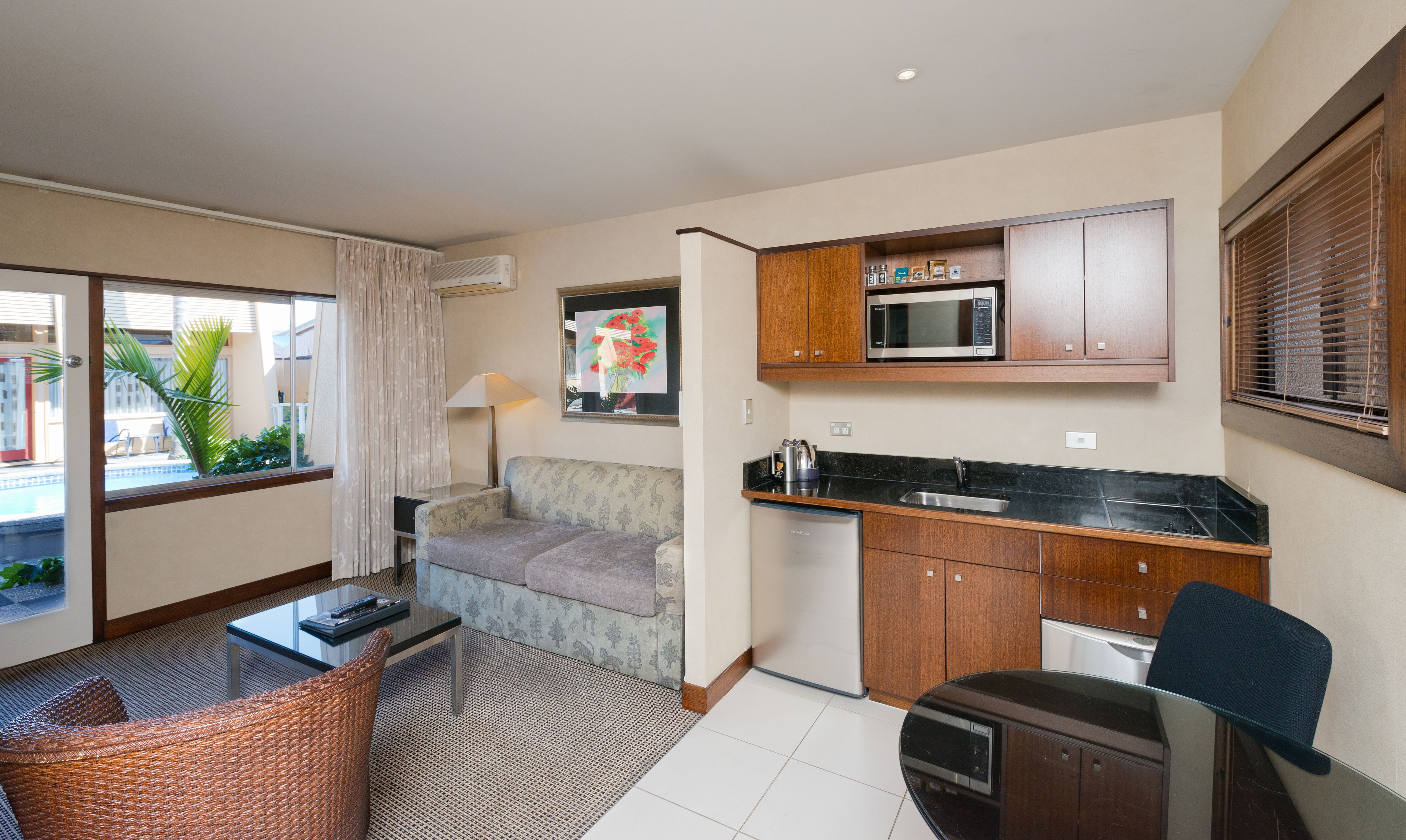 Habitación de hotel compacta con kitchenette, sofá, pequeña mesa de comedor, una silla y vistas al exterior.