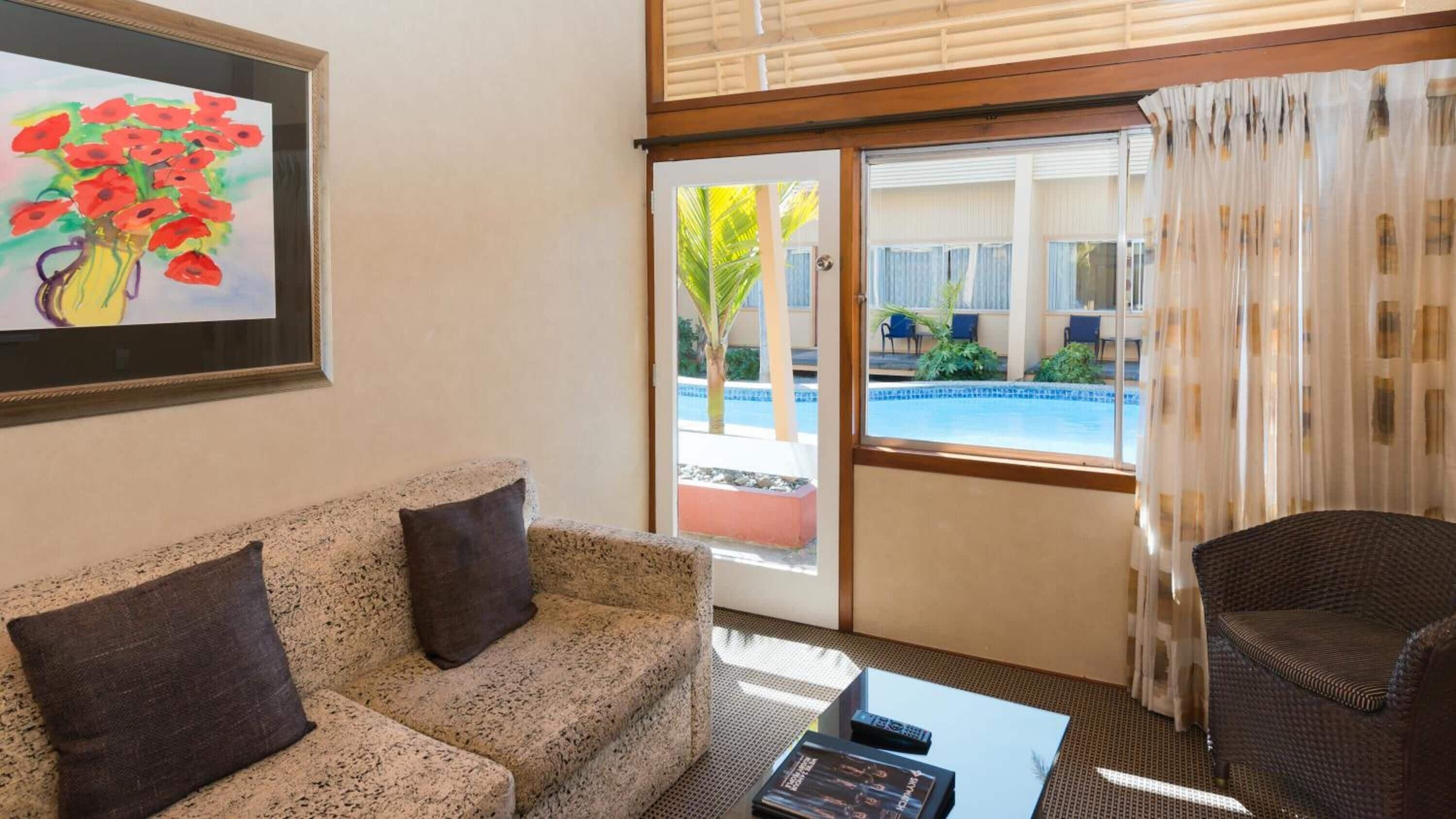 Un salon avec un canapé, une table basse et un fauteuil. Il y a un tableau au mur, une porte donnant sur un espace piscine et une fenêtre avec des rideaux.