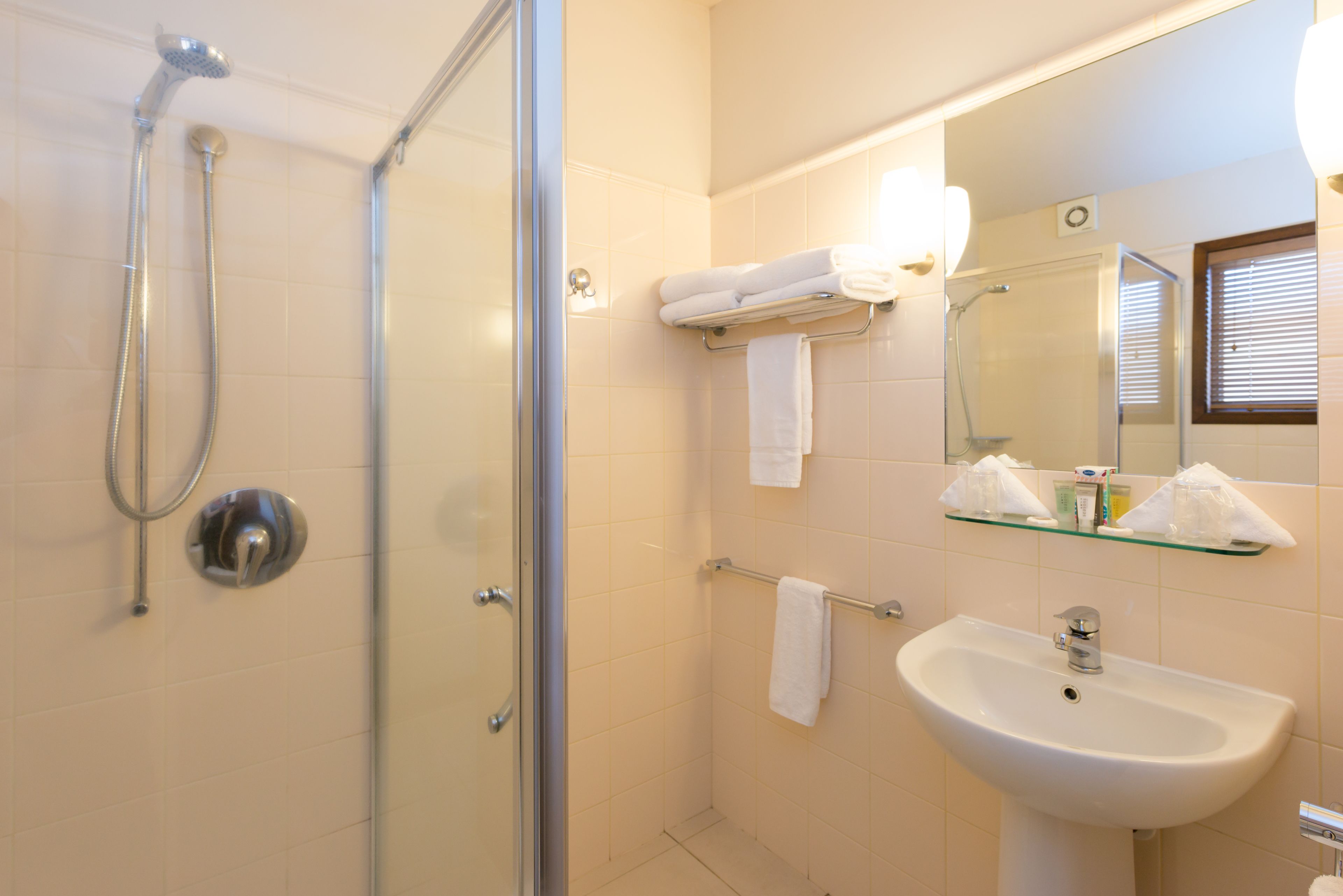 A bathroom with a glass-enclosed shower, a white sink, and a mirror with a glass shelf above it.