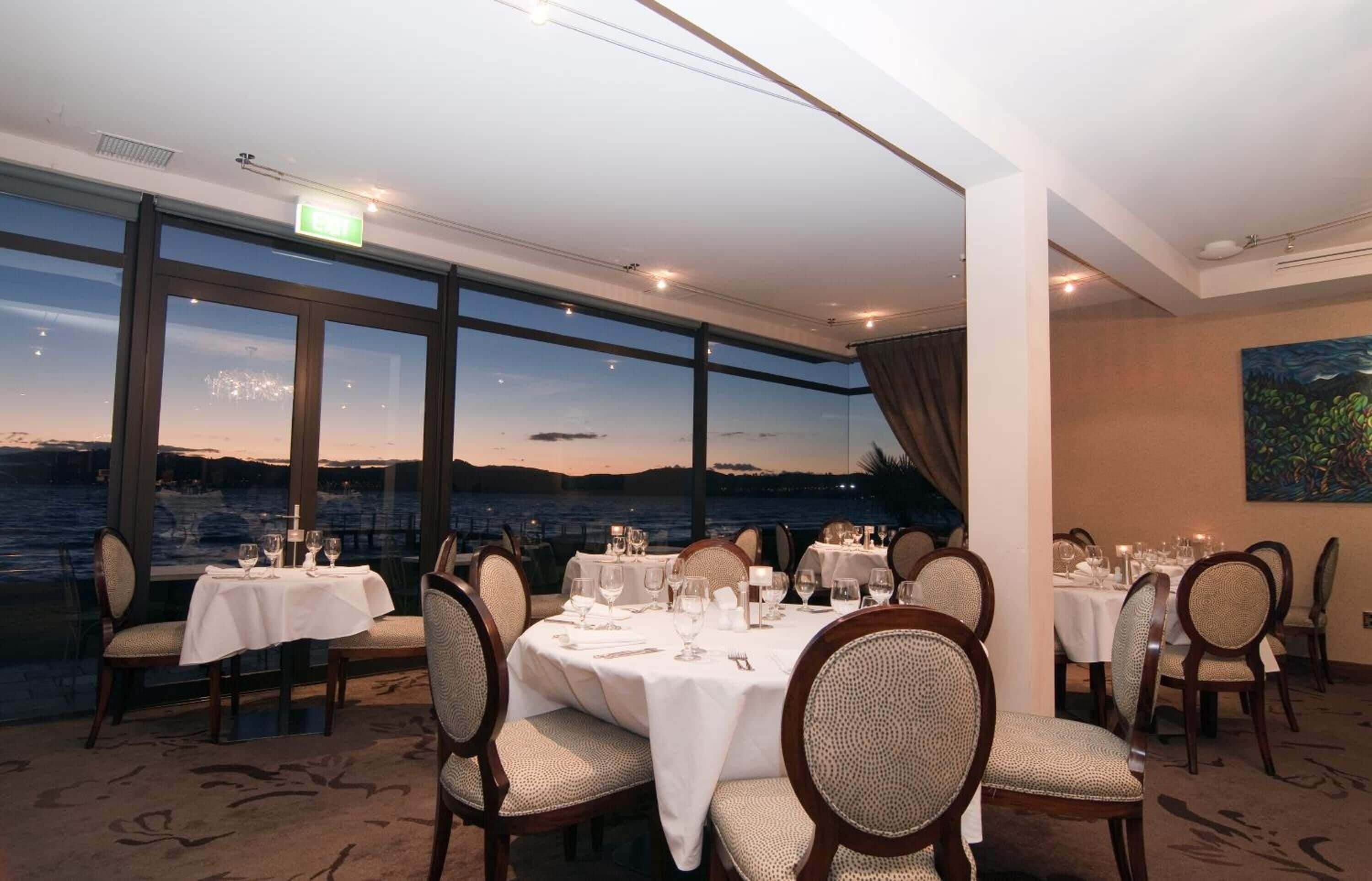 A dining area with tables set for a meal, large windows with a view of the sea, and a painting on the wall.