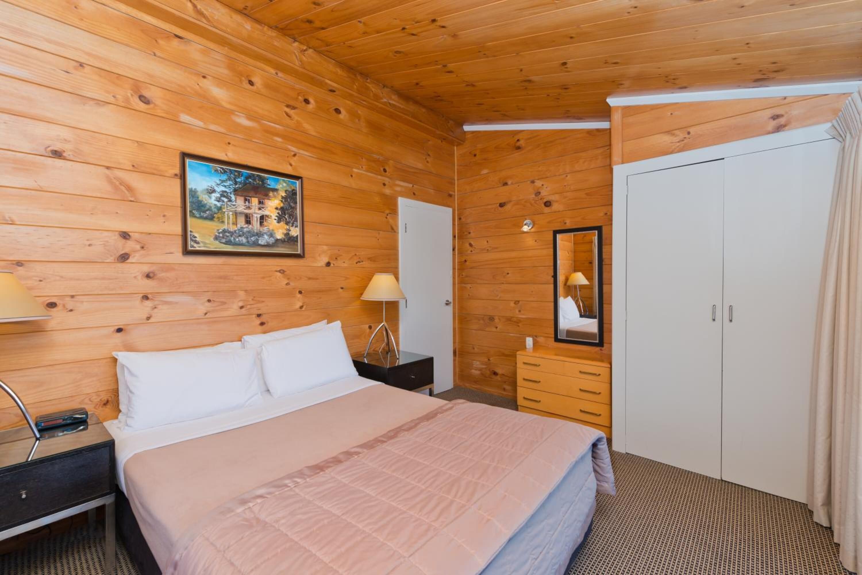 A wooden-paneled room with a bed, bedside table, lamp, dresser, and wardrobe.