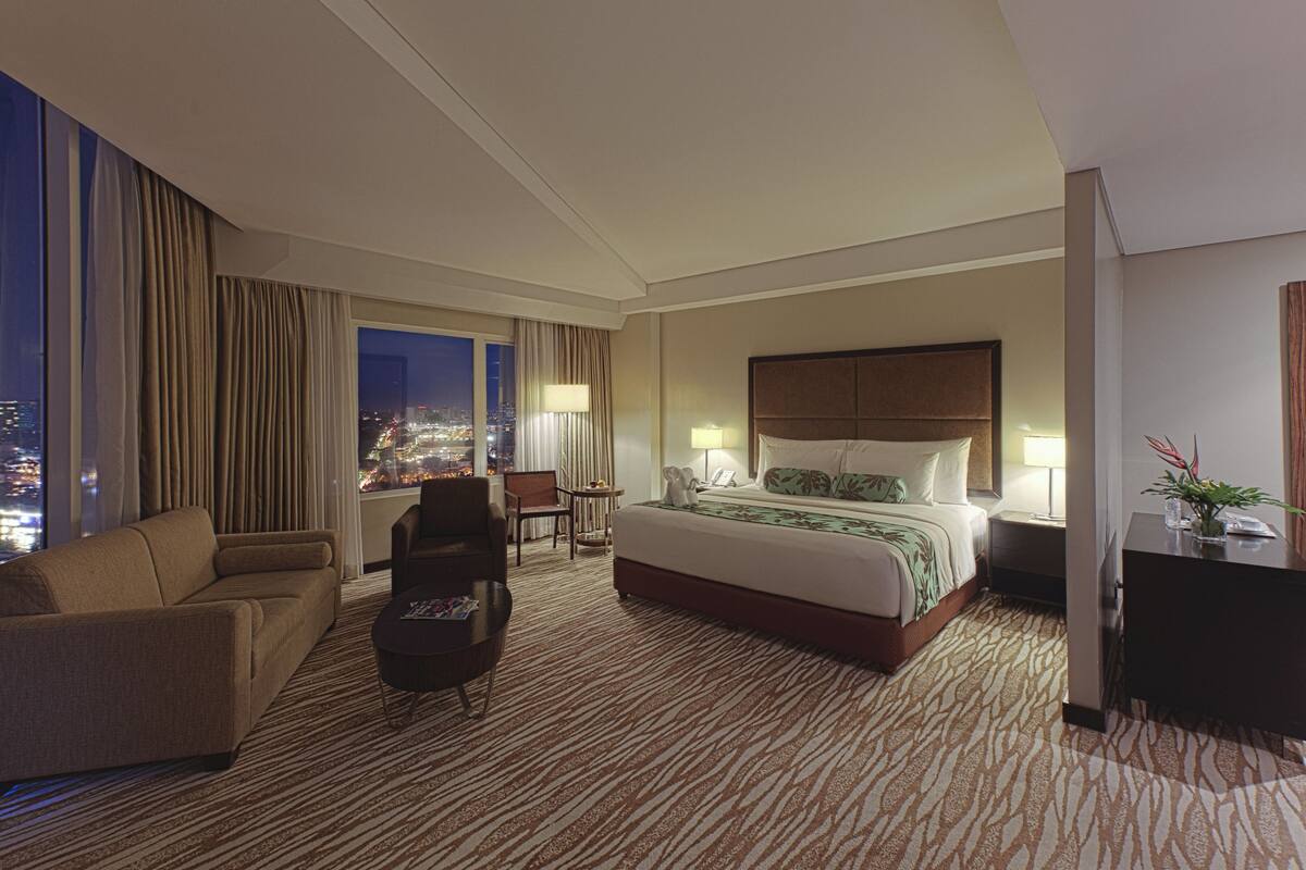 A hotel room with a large bed, a sofa, a desk, and a view of the city at night.